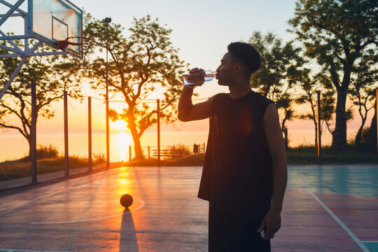 black man doing sports in morning, drinking water on basketball court on sunrise