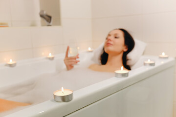 The girl lies in the bath with a glass of wine and candles. Selective focus on a candle. The girl is out of focus.