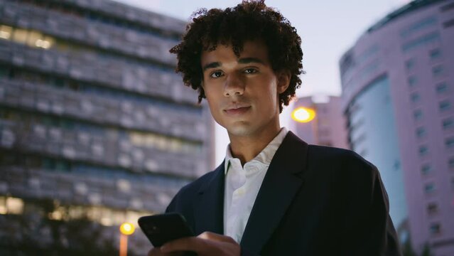 Formal suit guy holding phone at sunset city portrait. Young man looking camera