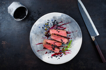 Gourmet barbecue dry aged angus roast beef steak with capers and rocket lettuce in amarena cherry sauce served as top view on a Nordic design plate with a Japanese santoku knife