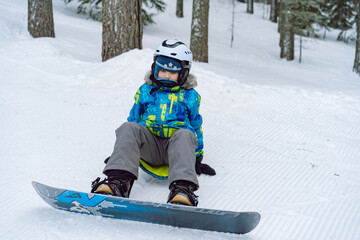 Fototapeta na wymiar little boy sitting on snow putting his feet in snowboard bindings adjusting straps. 