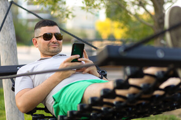 Happy young man resting on hammock with smartphone, reading news, chatting in social networks on sunny day