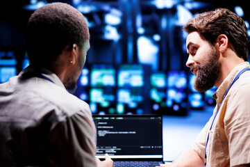 Coworkers updating blade servers, writing complex binary code scripts on laptop terminal. Employees...