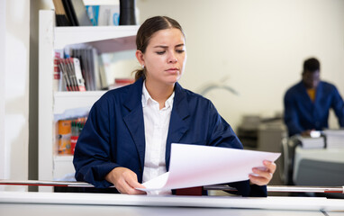 Attentive young female typographer in a blue uniform verifying printed sheet during work in the...