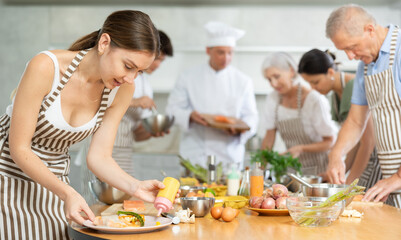 Interested engrossed young woman honing cooking skills during group culinary classes, garnishing...
