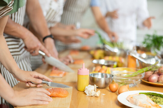 People in aprons standing at table with groceries and utensils during group cooking lesson, absorbed in cooking process, cutting fresh salmon following chef instructions. Cropped shot