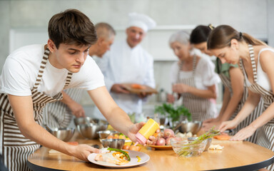 Enthusiastic young guy wearing striped apron honing cooking skills during group culinary classes, garnishing and seasoning finished dish with sauce before presentation