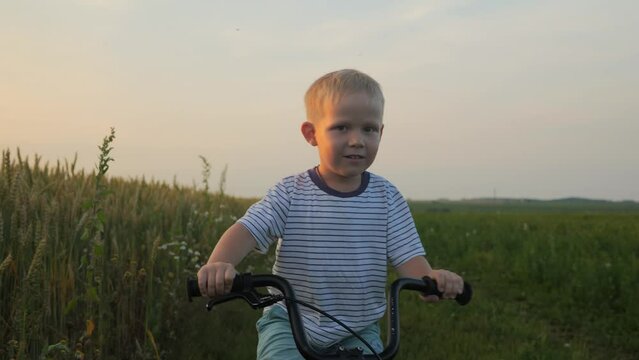 Portrait of a 5-year-old blonde boy in a striped T-shirt, who holds onto the handlebars of a bicycle and rides along a green field on a country road during sunset and looks into the camera. 4k footage