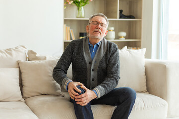 Knee pain arthritis body sick health care concept. Unhappy middle aged senior man suffering from knee ache sitting on sofa at home. Mature old senior grandfather touching leg feeling pain hurt in knee