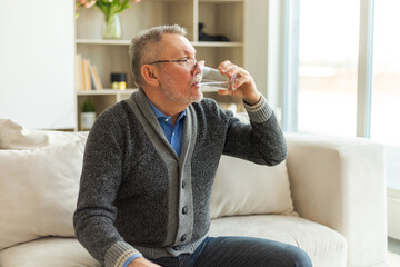 Senior man holding glass drinking fresh water at home. Mature old senior thirsty grandfather takes...
