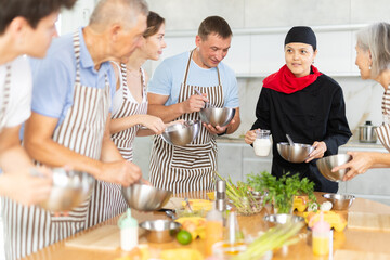 Positive young Asian woman professional chef conducting culinary courses, standing next to table with carafe of cream, imparting cooking skills to diverse group of people of different ages