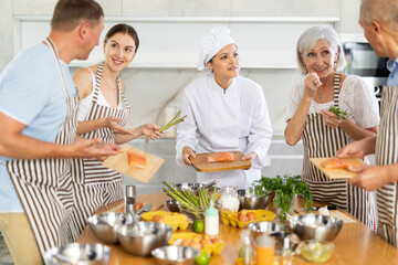Friendly skilled young Asian female chef giving culinary classes to group of men and women of different ages, teaching to cook salmon deliciously