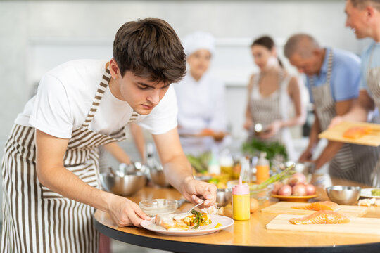 Attentive young male attendee of cooking course engaged in decoration of baked piece of salmon in the kitchen