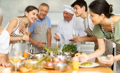 Positive friendly woman participating in group culinary masterclass led by professional chef, enjoying wine, engaging in cheerful conversations with other participants, and cooking together at table