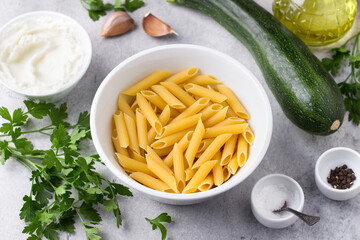 Penne pasta, zucchini, cream cheese, garlic, parsley, olive oil, salt, pepper for delicious vegetarian pasta on a gray textured background, top view