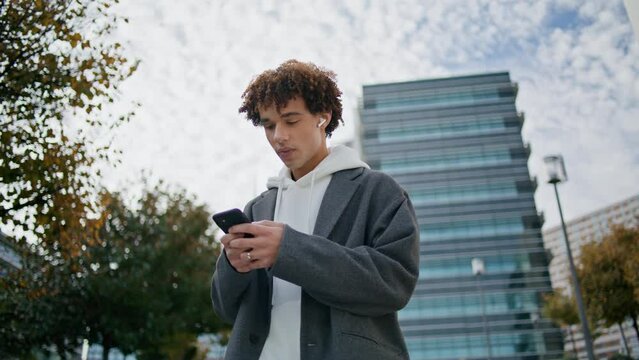 Curly teenager typing smartphone at street closeup. Young man listening music