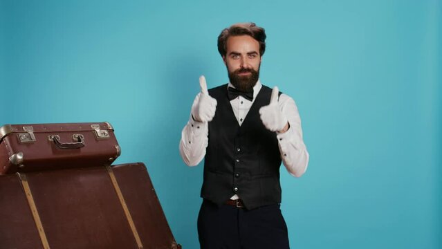 Hotel concierge shows thumbs up in studio, acting positive and optimistic against blue background. Joyful doorman does like and agreement symbol of acceptance, wearing formal attire.