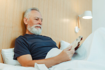 Gray haired elderly man lying in bed covered with blanket holding book, reading in stylish apartment