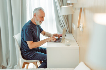 Smiling senior man sitting at desk in living room, hotel, using laptop, elderly copywriter typing