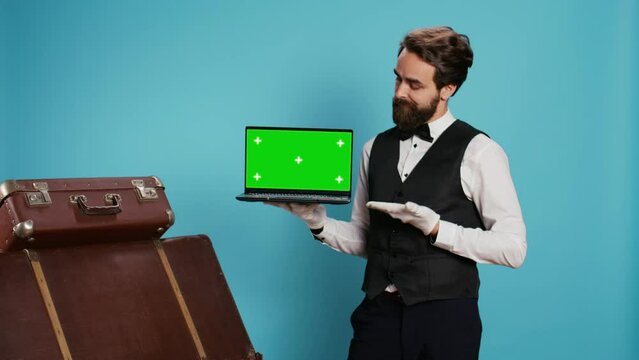 Classy bellhop presents greenscreen on laptop display in studio while he wears gloves next to pile of luggage. Experienced doorman working at luxurious hotel, showing isolated mockup.