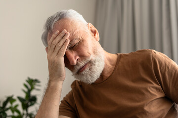 Portrait of tired senior man with closed eyes touching head, having headache in living room