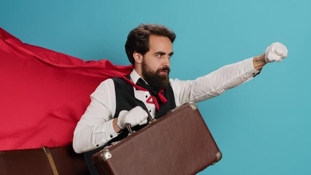 Hotel bellhop acting as superhuman with cape, offering to help people with suitcase luggage in studio. Young adult doorman presenting comic hero character with red fabric, feeling determined.