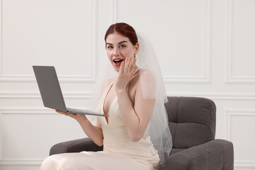 Fototapeta premium Surprised bride with laptop sitting in armchair indoors