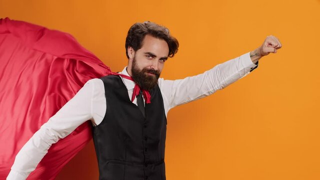 Elegant butler with red cape posing on camera while he acts like superhuman at luxury restaurant, yellow studio. Young man catering worker serving food and drinks, wearing hero mantle.
