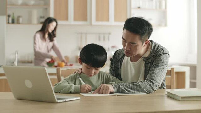 young asian father helping son with homework at home while mother preparing meal in kitchen