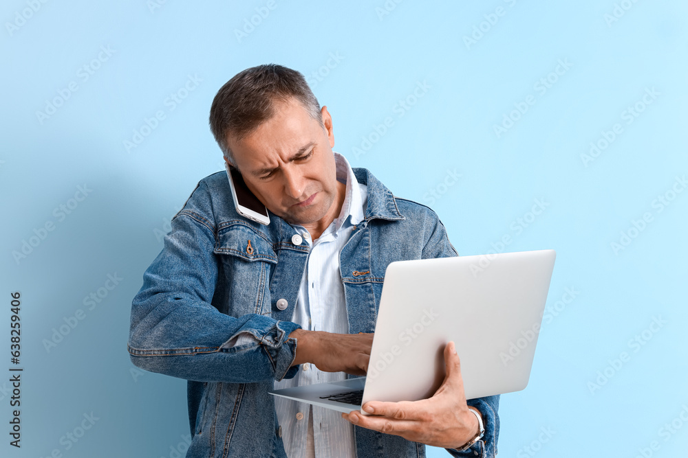 Wall mural stressed mature man with laptop talking by mobile phone on blue background. deadline concept