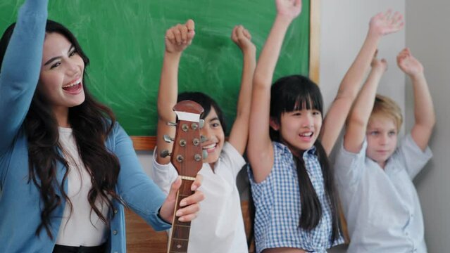 Teacher female with guitar and sitting lined up with students in front of blackboard and raise hand in music subject at elementary school