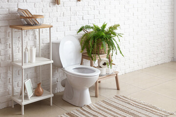 Interior of light restroom with ceramic toilet bowl, shelving unit and houseplant near white brick wall