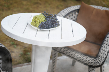 A dish with grapes stands on a white table, in the open air, in a summer cottage. - obrazy, fototapety, plakaty