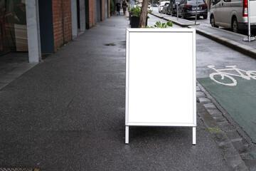 Background texture of a blank white mockup street board template placed outdoor on the pedestrian walkway. Empty advertisement sandwich easel panel on side of urban road. Copy space for your design.