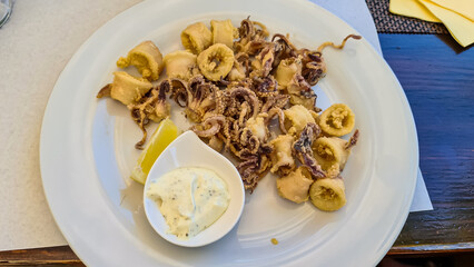 A plate of fried calamari served with a piece of lemon and garlic sauce. The dish is prepared on a white, round plate and located on a wooden table. Mediterranean cuisine. Frutti di mare.