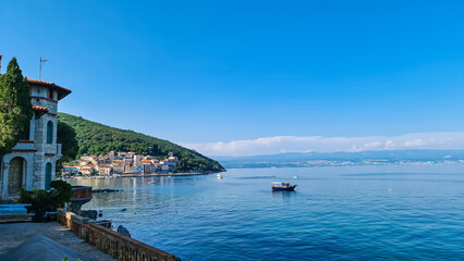A panoramic view of the shore along Moscenicka Draga, Croatia. There is a small town located on the shore of the Mediterranean Sea. Few boats crossing the calm sea. Green hills in the back. Sunny day.