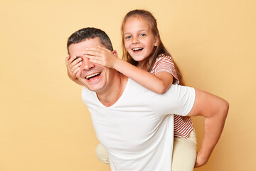Joyful laughing female kid piggybacking on father isolated over beige background covering dad's eyes with hands laughing having fun playing together.