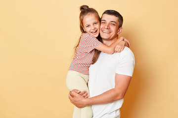 Smiling brunette man holding his little daughter, child hugging her father family looking at camera with happy faces standing in casual clothing isolated over beige background.