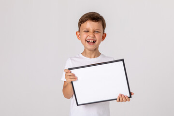 Fototapeta premium portrait of happy laughing baby boy holding white drawing Board on white background, young boy is holding a drawing board in her hands. white isolated background. Mock up. Copyspace.