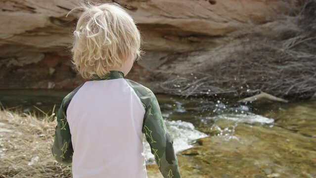 Over the shoulder 3 year old boy throws rock into river, stream, Escalante Utah