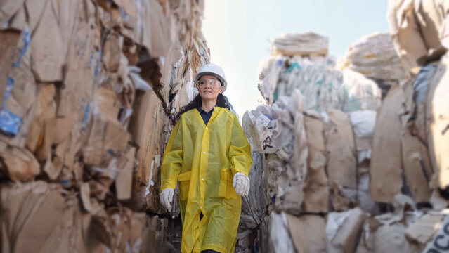 Long-haired woman walks past pile of trash at waste sorting plant. Manager in hardhat explores warehouse with paper bales for recycling of unsorted trash