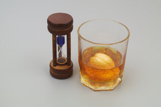 Hourglass and human brain drowned in alcohol drink on gray background.	