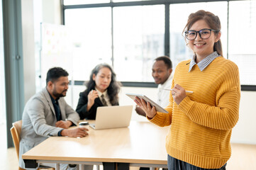 A smiling Asian businesswoman in glasses standing with a tablet in her hand in a meeting room