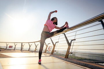 Woman in tracksuit streches muscles on legs on footbridge. Active exercise and healthy lifestyle in public place low angle shot