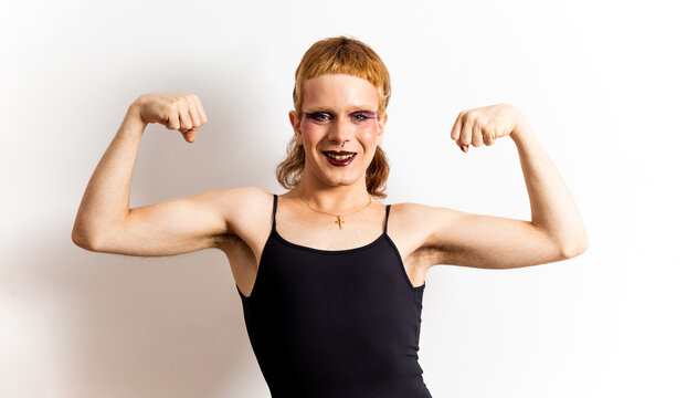 A red-haired non-binary person wearing makeup in a black T-shirt poses on a white background while showing off both biceps. Concept of support for the LGBTI collective, strength for LGBT people.