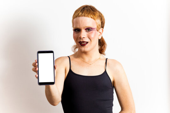A non-binary redhead in makeup poses in a black T-shirt on a white background while showing a mobile phone with a white screen. Concept of non-binary people, mobile advertising.