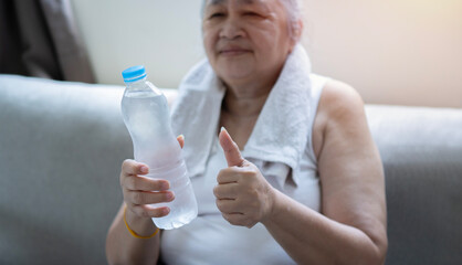 Portrait of a older woman show thump up and a bottled water while sitting on sofa at home.