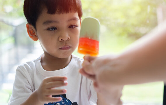 Picture of cute Asian boy looking and taking ice cream from mother 's hand on blurred background of nature.
