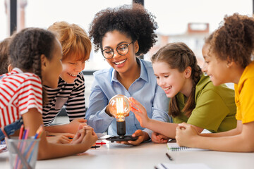 Multiethnic teacher and kids studying electricity and incandescent light bulb at physics lesson. - obrazy, fototapety, plakaty