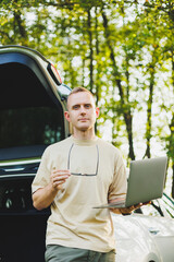 Cheerful traveler, young man sitting in open trunk of car, working with laptop, outdoors, concept of business, travel, vacation